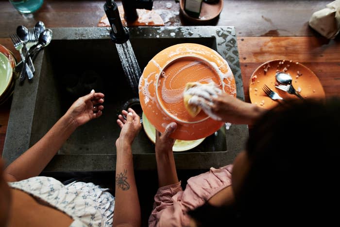 Two people wash dishes in a kitchen sink; one holds a soapy plate, while the other assists. Various kitchen utensils are scattered around