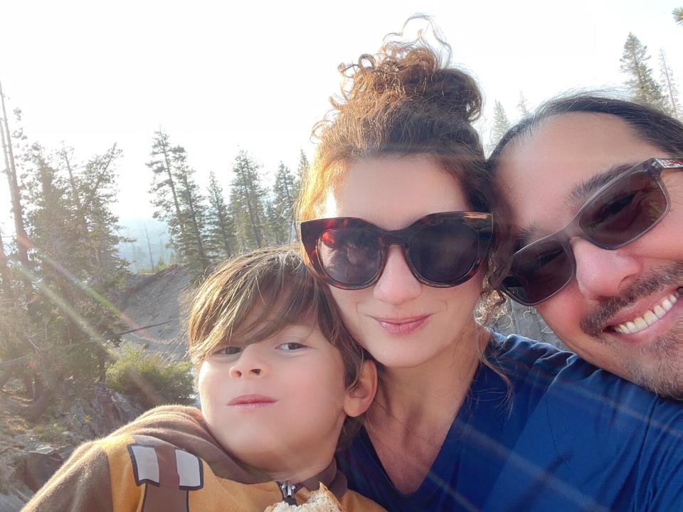 A young boy with a woman and a man who both wear sunglasses, outside with tall trees in the distance