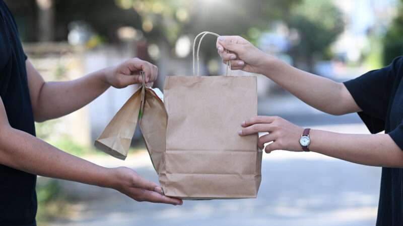 A delivery driver hands off a paper bag of takeout food to a customer.