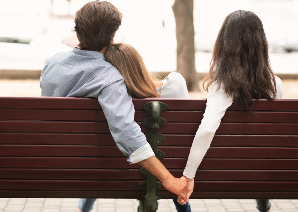 A man and two women sitting on a bench, with the man holding hands secretly with the woman beside him