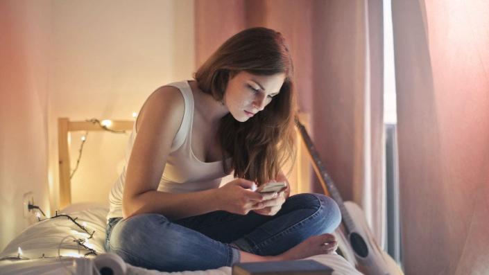 Woman sits in bedroom while texting on her phone