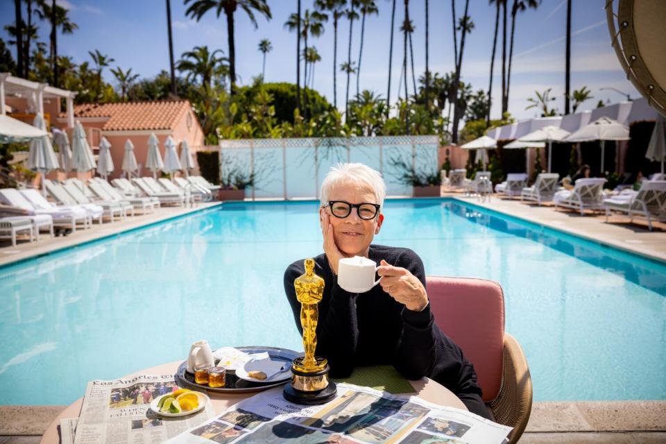 A woman in glasses holds a cup with latte foam next to a table with her Oscar on it.