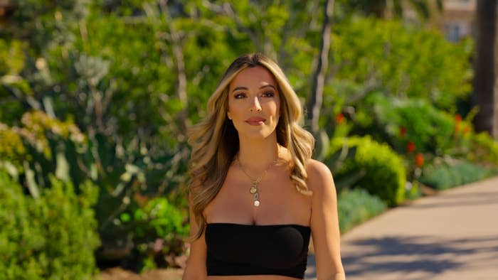 Alex in a black strapless top with pendant necklace, standing on a garden path