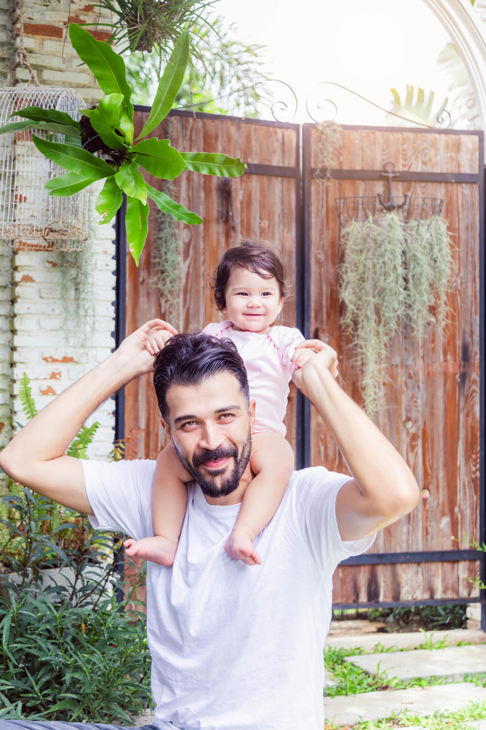 A man with a beard wearing a white shirt smiles while giving a piggyback ride to a cheerful toddler girl in a garden setting