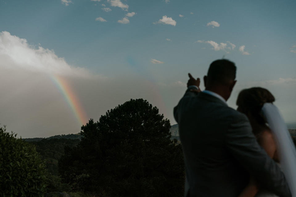 A silhouette shot of the bride and groom outdoors. The groom has his right arm wrapped around the bride as he points to a rainbow in the distance.&nbsp; (Photo: <a href="https://www.jamesday.com.au/" target="_blank">James Day Photography</a>)