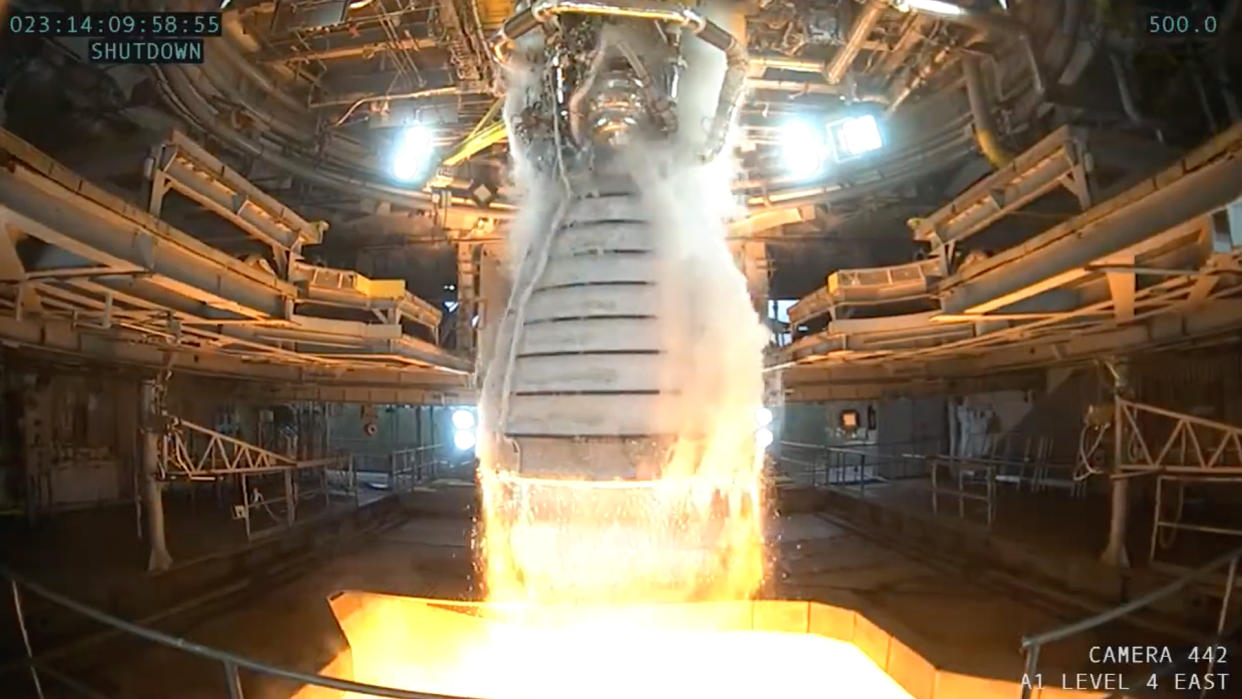  Close-up of a rocket engine with steam pouring out the sides and fire at the bottom. surrounding is a large test stand. 