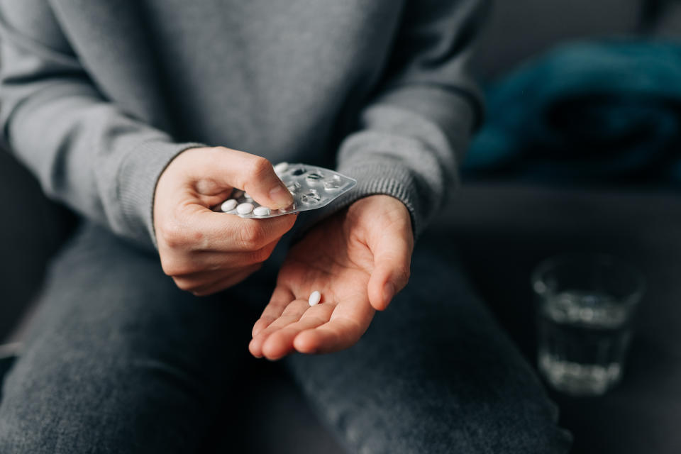 Person holding a blister pack of pills with one pill in hand, sitting with a glass of water nearby