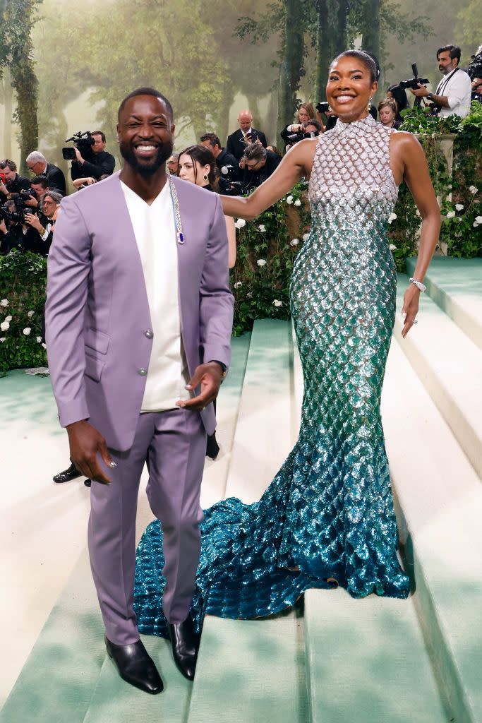 Two smiling individuals on a green carpet, one in a purple suit, the other in a shimmering gown