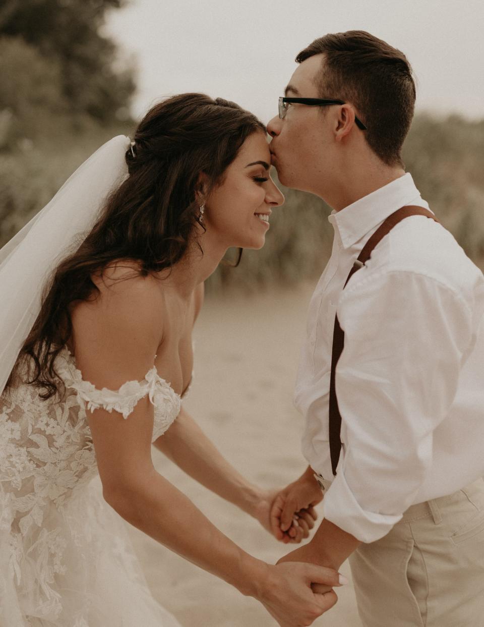 A bride kneels down as her brother kisses her forehead on a beach.