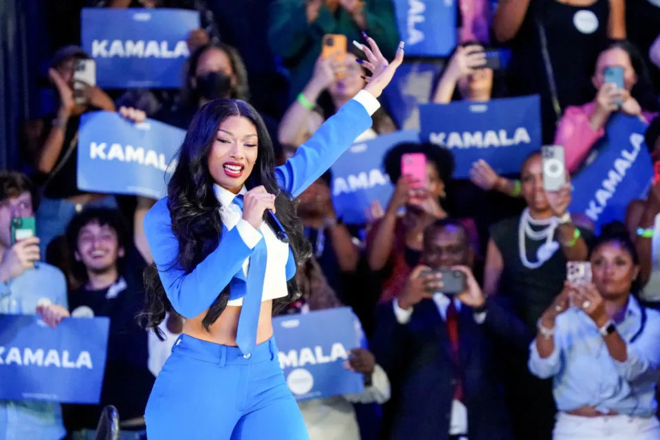 Megan Thee Stallion performs on stage, wearing a stylish blue outfit, surrounded by fans holding signs that say "KAMALA."