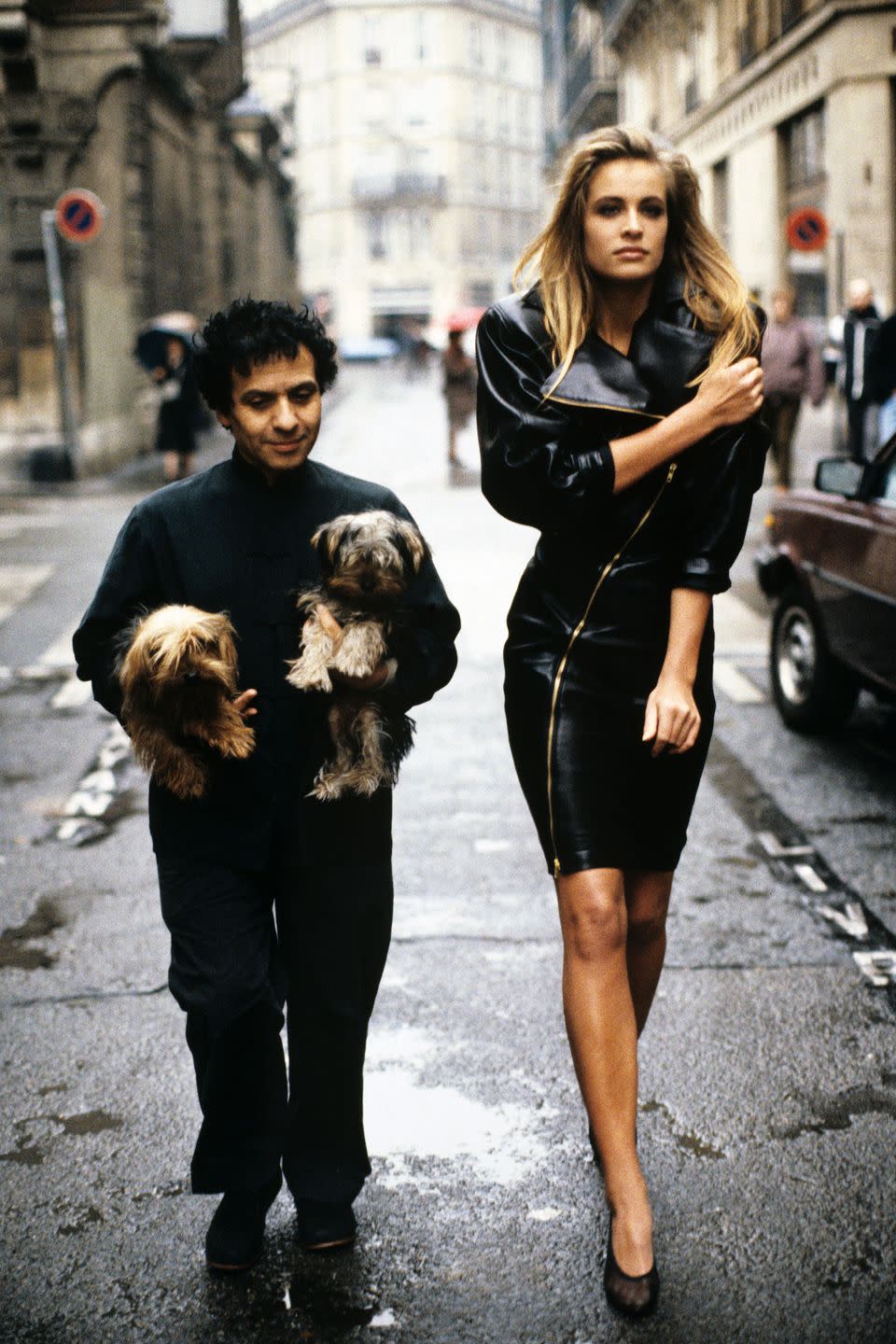 <p><em>Azzedine Alaia holding his two Yorkshire terriers, Patapouf and Wabo, walking in Paris street with model Frederique who wears one of his creations, a black leather zippered dress, 1986</em></p>