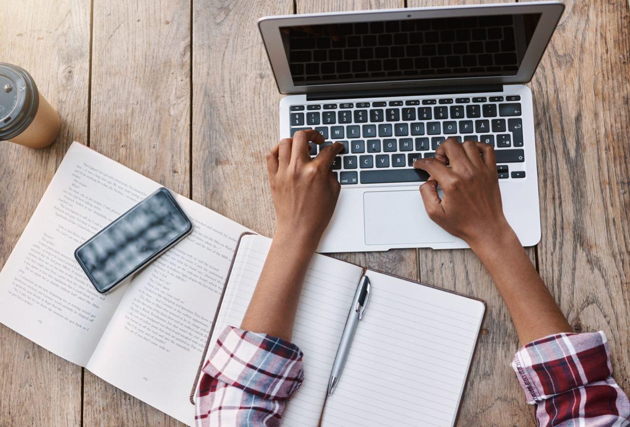 An image of a student typing on their laptop with a notebook next to them.