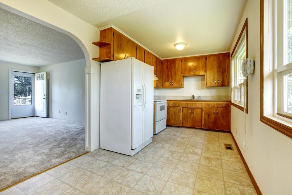 View of the empty living room from a small kitchen with concrete floor, white appliances and wooden cabinets.