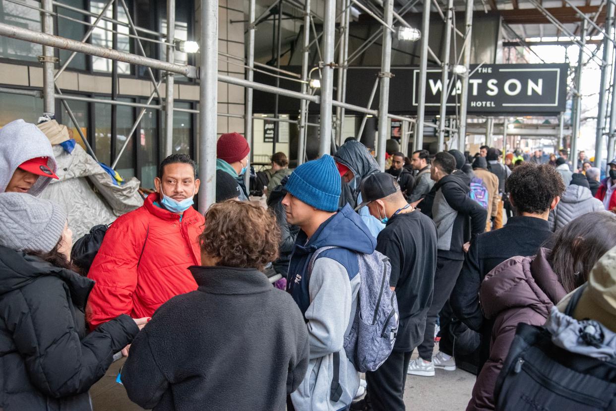 A large number of migrants in winter clothes, some wearing masks, in conversation outside the Watson Hotel, which is covered in scaffolding. 