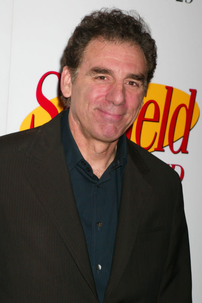 A person smiles at an event with a background featuring the "Seinfeld" logo. They are wearing a dark suit and a navy shirt