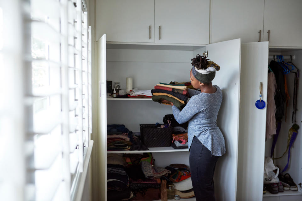 A person wearing a long-sleeve shirt and headband is organizing clothing in a closet. The closet has several shelves filled with folded clothes