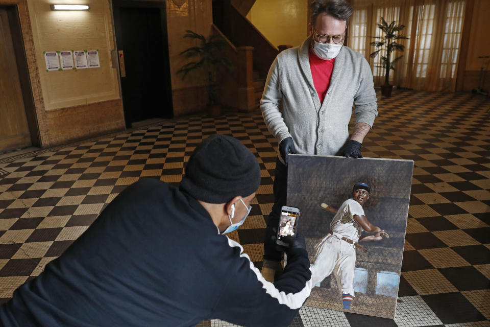 In this Wednesday, April 29, 2020, photo, delivery man Pablo Alfaro, left, takes a cell phone photograph of baseball artist Graig Kreindler's portrait of Josh Gibson while making a drop-off, as Kreindler holds his painting during a photos session in the lobby of his residence in the Brooklyn borough of New York. (AP Photo/Kathy Willens)