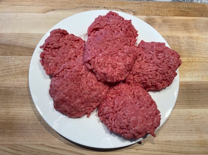 Six raw hamburger patties on a white plate on a wooden cutting board