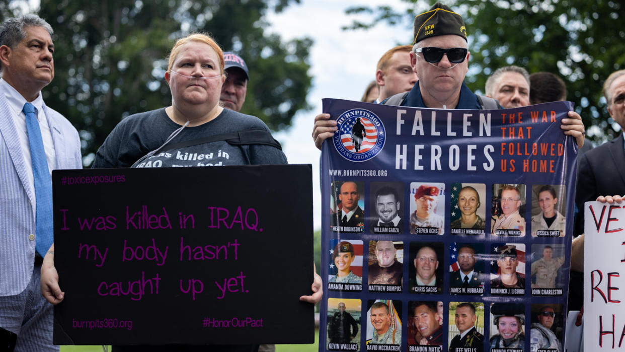 Veterans and supporters of the Honoring Our PACT Act stand near a podium holding signs that read: I was killed in IRAQ. My body hasn't caught up yet and Fallen heroes: The war that followed us home.