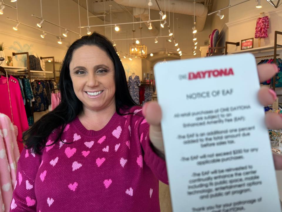 Brooke Kells, store manager and buyer at the Pink Narcissus women's resort wear shop at One Daytona, holds up a sign explaining the 1% "Enhanced Amenity Fee (EAF)" that stores and restaurants at the NASCAR-owned entertainment/retail center charge customers on Thursday, Jan. 18, 2024. Most customers don't notice it, but those that do "don't like it. They don't understand it," she said.