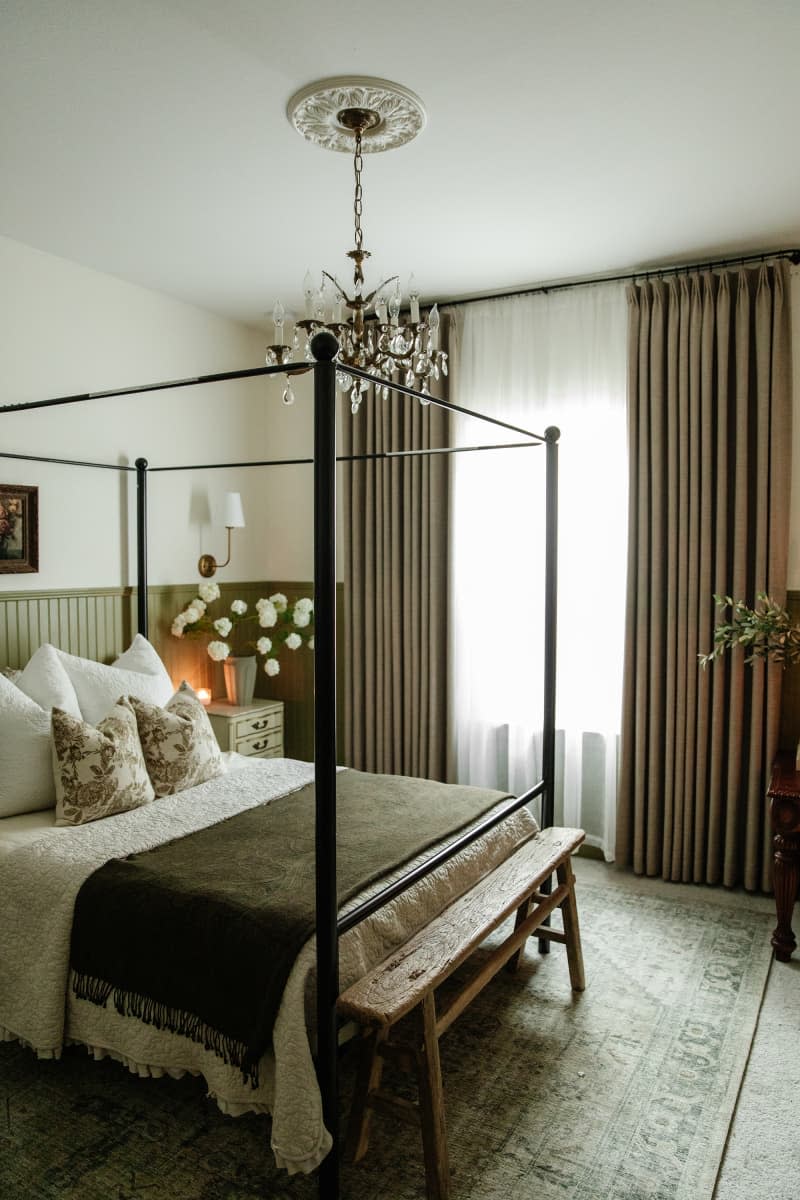 Bedroom with natural hues, green and white walls, canopy bed, chandelier, large windows with floor to ceiling curtains, white flowers in vase