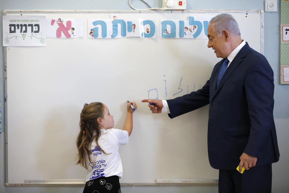 Israeli Prime Minister Benjamin Netanyahu stands next to a student as she writes on a board during a ceremony opening the new school year, Sunday, Sept. 1, 2019, in the West Bank Israeli settlement of Elkana. Speaking Sunday at the ceremony, Netanyahu reaffirmed his pledge to impose Israeli sovereignty on Israeli-occupied West Bank settlements. The writing in Hebrew on the board reads, "Hello." (Amir Cohen/Pool Photo via AP)
