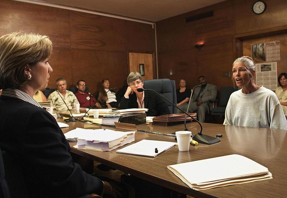 a parole board member sits at a table and listen to leslie van houten, who wears a gray sweatshirt and speaks as several people watch from seats in the background