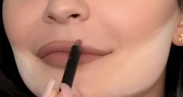 A person is having their lipstick applied with a makeup brush. Their face shows partially applied makeup, including contouring powder