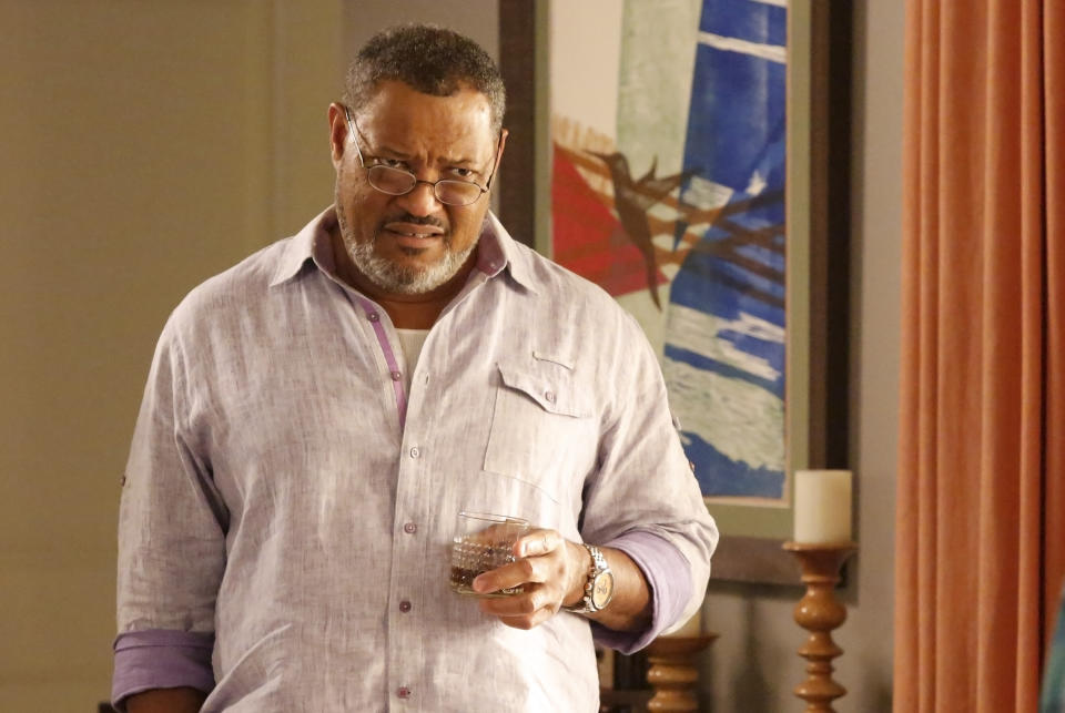 Laurence Fishburne stands indoors, holding a drink and wearing a casual light-colored shirt. Artwork and a curtain are visible in the background