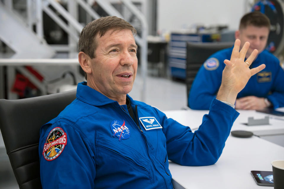 a man wearing a blue suit sits in a white room with black furniture.  his elbow is resting on a table.