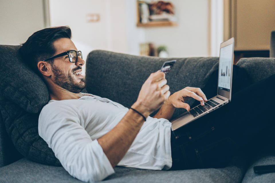 Man smiling at credit card while using laptop, reflecting on online dating expenses