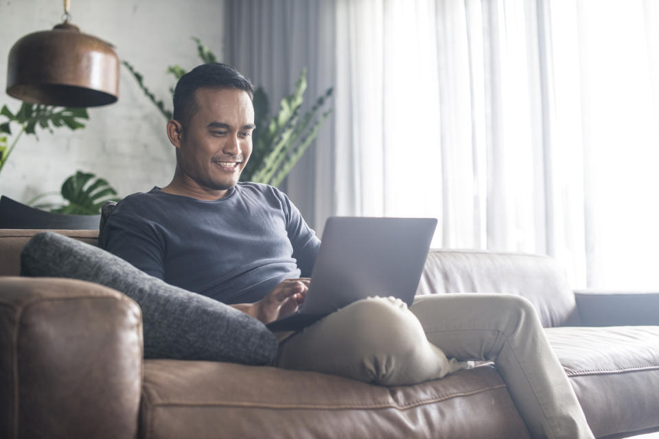 Smiling man sitting on couch, typing on laptop.