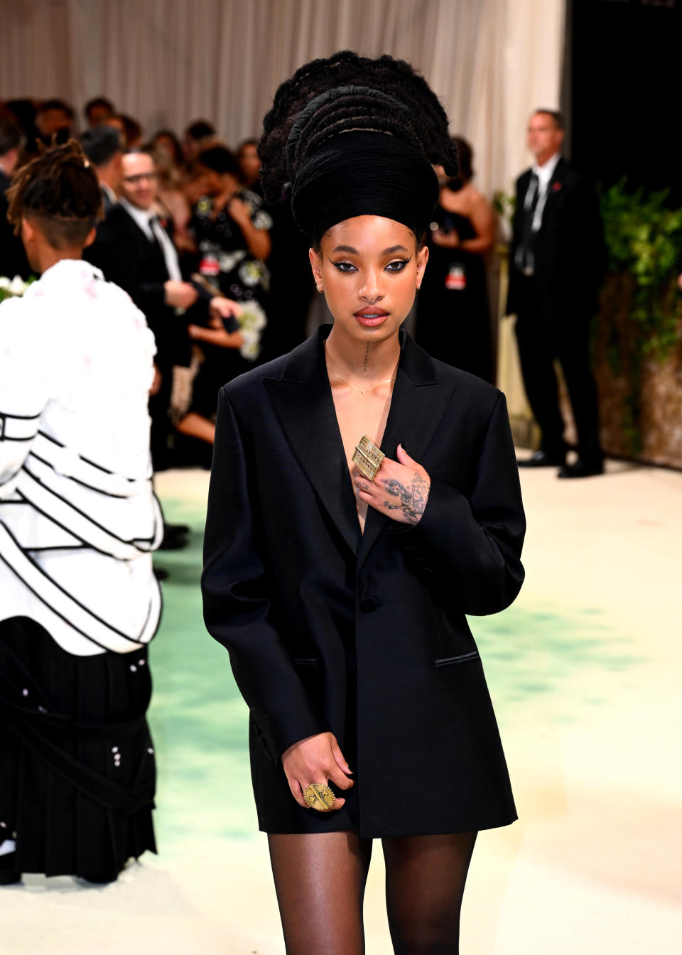 Willow Smith wearing a black oversized blazer and statement ring on the red carpet at a celebrity event, with attendees in the background