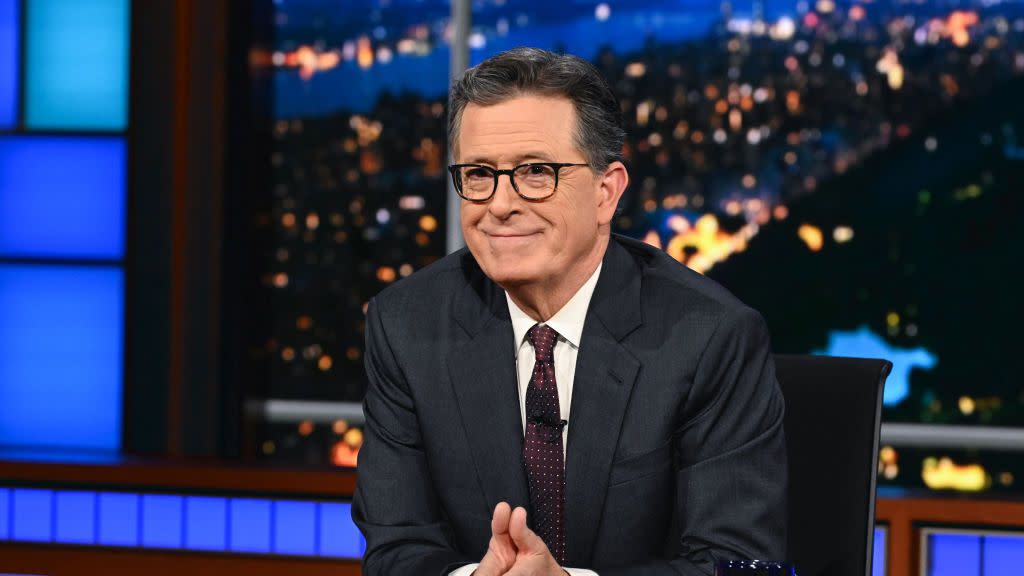 stephen colbert sitting at his desk with his palms together