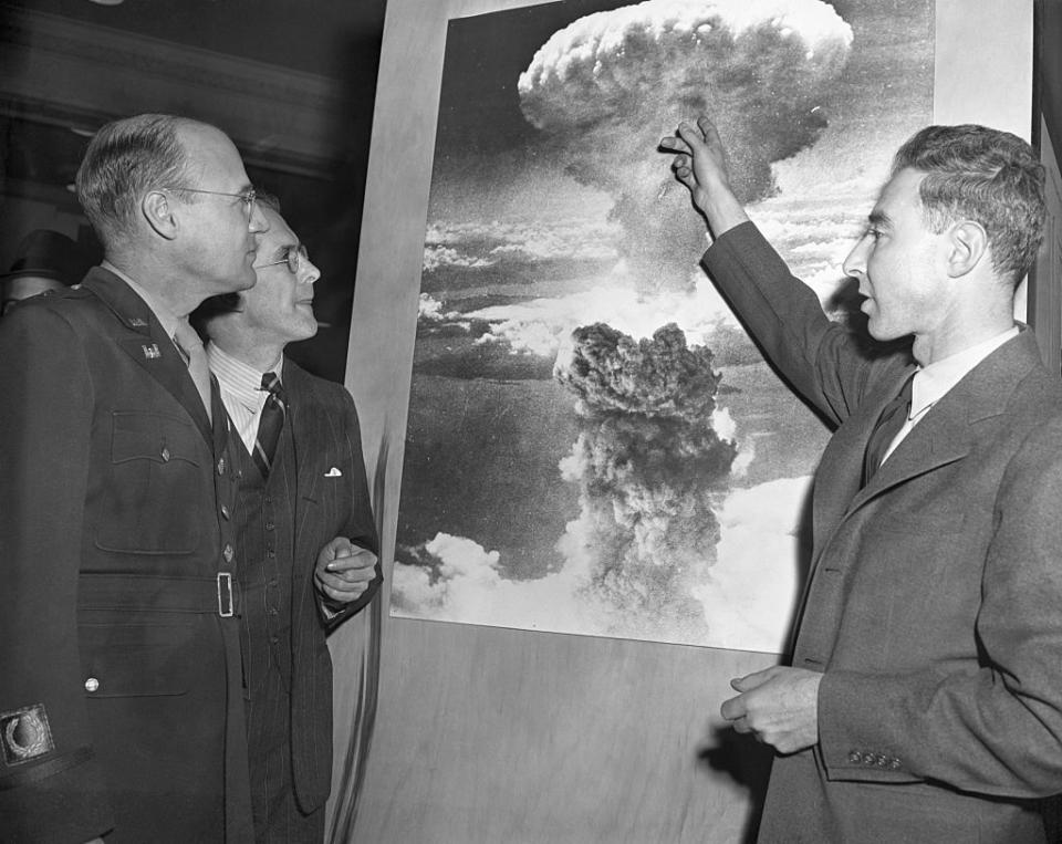 a man in a military uniform and a man in a suit and tie listen as j robert oppenheimer, also wearing a suit and tie, points at a photo of the atomic bomb detonation
