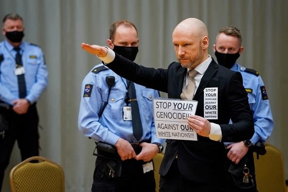 Anders Behring Breivik, in a suit, performs a Nazi salute while holding signs. Three uniformed police officers are present in the background