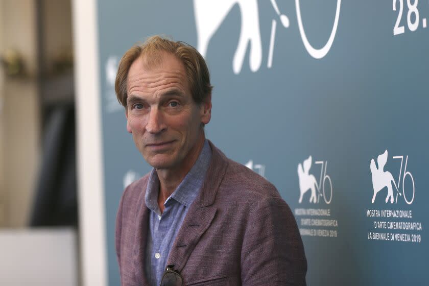 A man in a brown blazer and blue button up shirt leans forward to pose for pictures