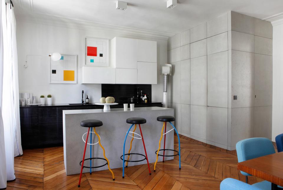 The cabinets and island are IKEA covered in concrete. The colorful stools are Duplex by Javier Mariscal, edited by Ediciones de Diseno, from 1981. The floor lamp is called Shogun Terra by Mario Botta for Artemide  and the wall light is Serge Mouille. The serigraphs are by Gottfried Honegger from 2015 and the marble bowl is by Angelo Mangiarotti.