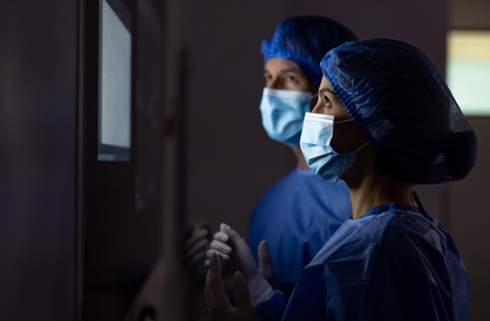 Two healthcare workers wearing surgical gowns, gloves, masks, and hair caps look at a screen intently. The context suggests a work-related setting