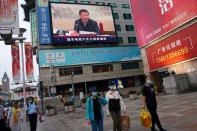 People hold shopping bags as they walk under a giant screen showing a news footage of Chinese President Xi Jinping, at a shopping area in Beijing