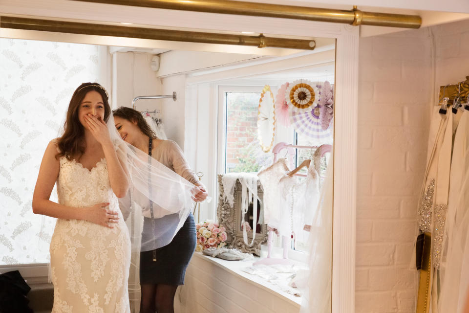 Bride in lace gown smiling as person adjusts her veil by a mirror, surrounded by wedding dresses