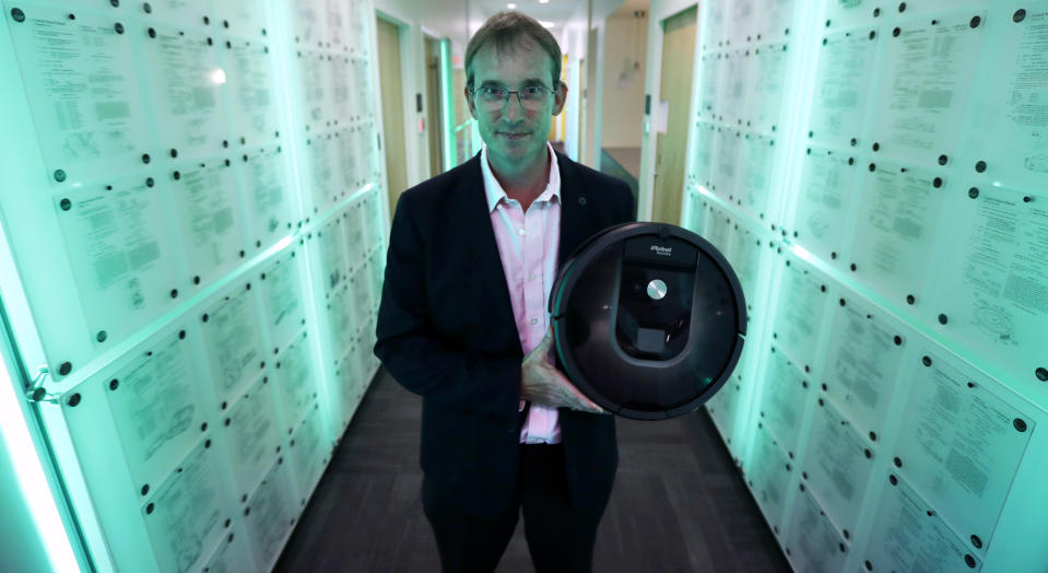 FILE - In this Thursday, Aug. 25, 2016, file photo, iRobot co-founder and CEO Colin Angle is illuminated in blue-green light while holding a Roomba vacuum in a hallway decorated in patents the company owns, at their headquarters in Bedford, Mass. Building a robot lawn mower seemed the logical next step for iRobot, which invented the pioneering robotic vacuum Roomba. Terra, the company’s long-awaited first lawn mower, is being publicly unveiled Wednesday, Jan. 30, 2019. (AP Photo/Charles Krupa, File)