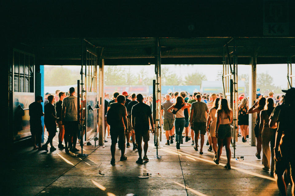 People walking toward an outdoor event through large open doors, with sunlight streaming in from the outside. The scene is crowded and lively