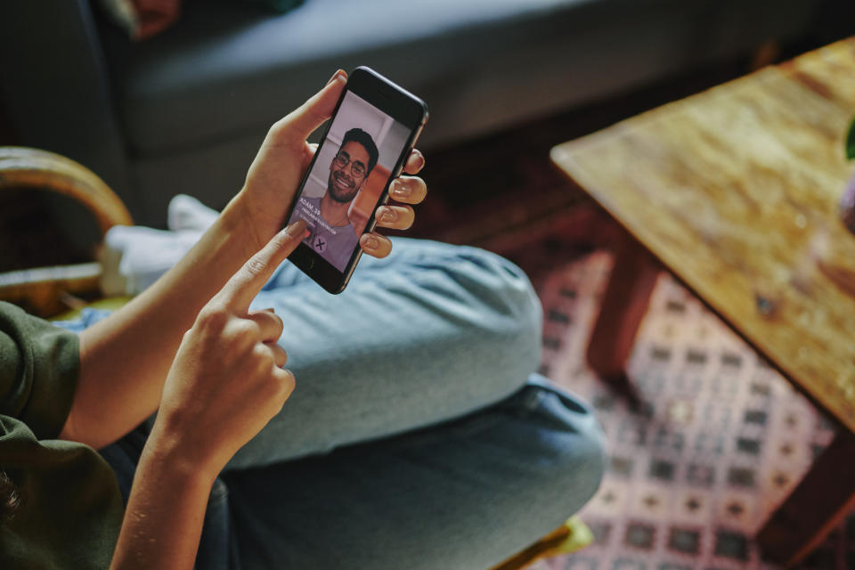 A person sits on a chair, looking at and touching a smartphone screen displaying a man's face during a video call