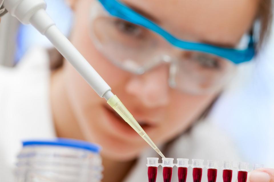 A pharmaceutical lab technician using a pipette to place red liquid samples into a row of test tubes. 