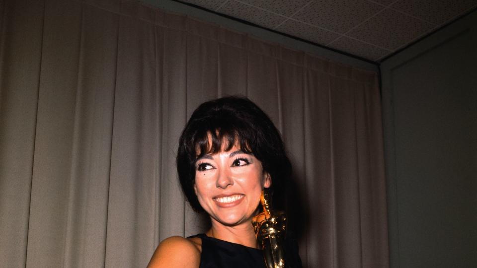 rita moreno smiles and holds an academy award statuette, she wears a black dress and long black gloves