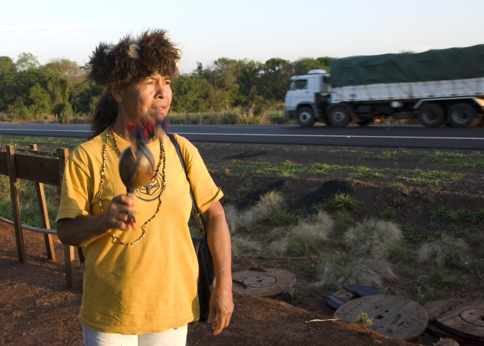 Damiana Cavanha stands by the side of a Brazilian road, a blue-feathered maraca made from a pumpkin gourd in one hand, and starts to sing. The ground is littered with trash; behind her stand huts constructed from corrugated iron, plastic sheeting and tarpaulin. Trucks thunder past; the noise drowns out her invocations. (Photo by Survival International)