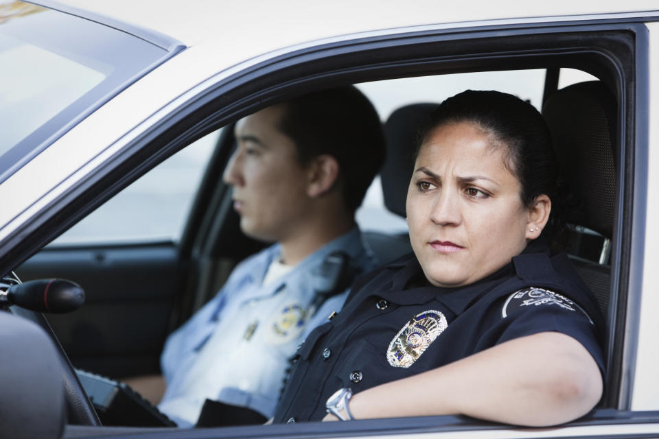 Two police officers sit in a patrol car, one looking directly ahead, the other to the side. Both are in uniform, focused on their surroundings