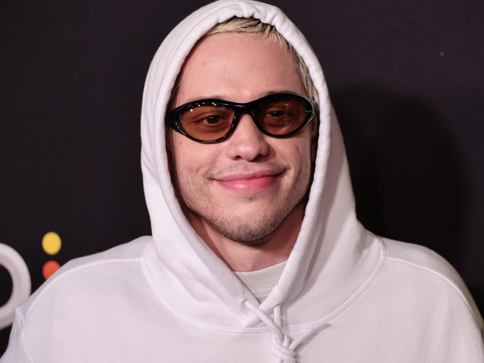 pete davidson wearing yellow tinted sunglasses and a white hoodie, pulled over his head and smiling