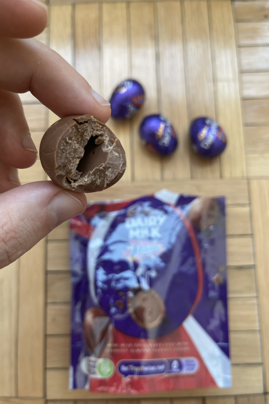 A hand holding a half-eaten chocolate with more chocolates and their purple packaging in the background